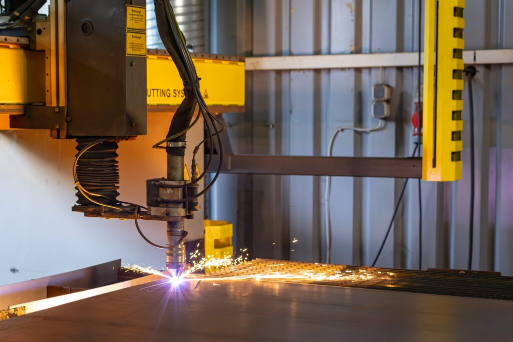 Das Bild zeigt eine industrielle Schneidemaschine, die gerade ein Metallstück bearbeitet. Aus der Schneidstelle sprühen glühende Funken in einem leuchtenden orange-gelben Farbton, die durch die hohe Hitze des Schneideprozesses entstehen. Der Schneidekopf ist über mehrere Kabel und Schläuche mit der Maschine verbunden und wird von einer gelben Struktur gehalten. Die Umgebung deutet auf eine Werkshalle hin, mit Kabeln und mechanischen Teilen im Hintergrund. Das Bild vermittelt den Eindruck von präziser, automatisierter Metallbearbeitung und zeigt einen Moment intensiver Energie beim Schneiden.