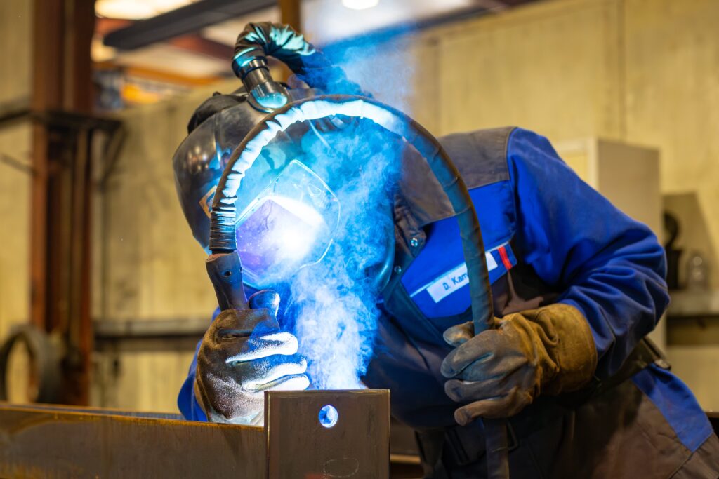 Das Bild zeigt einen Schweißer in voller Schutzkleidung, der konzentriert an einem Werkstück arbeitet. Die Person trägt eine dunkelblaue Schweißermaske und dicke Schutzhandschuhe, während sie einen Schweißbrenner hält. Ein intensives, blaues Licht leuchtet von der Schweißstelle, wodurch die Umgebung dramatisch erhellt wird und eine Wolke aus Schweißrauch aufsteigt. Der Name „D. Kamp“ ist auf dem Namensschild der Arbeitskleidung zu sehen. Die Szene vermittelt den Eindruck von Präzision und Konzentration und zeigt die raue, technische Umgebung eines metallverarbeitenden Betriebs.