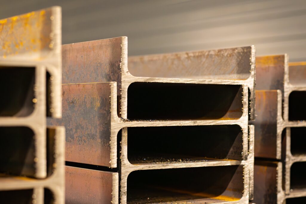 Das Bild zeigt eine Nahaufnahme von mehreren robusten Stahlträgern mit einer H-Profilform, die aufeinandergestapelt oder nebeneinander angeordnet sind. Die Kanten der Stahlträger wirken scharf und zeigen deutliche Spuren von Bearbeitung, wie leichte Schleifspuren und winzige Metallspäne, die an den Oberflächen haften. Die Oberfläche der Träger ist teilweise mit einer rötlich-braunen Patina überzogen, was auf eine leichte Oxidation oder Rückstände von der Herstellung hindeuten könnte. Das H-Profil dieser Stahlträger verleiht ihnen eine hohe strukturelle Stabilität, was für Bau- oder Maschinenbauprojekte wichtig ist. Das Bild vermittelt die industrielle Stärke und Solidität des Materials und zeigt die Rohheit und Stabilität, die bei der Verarbeitung von Stahl im Bauwesen gefragt ist.