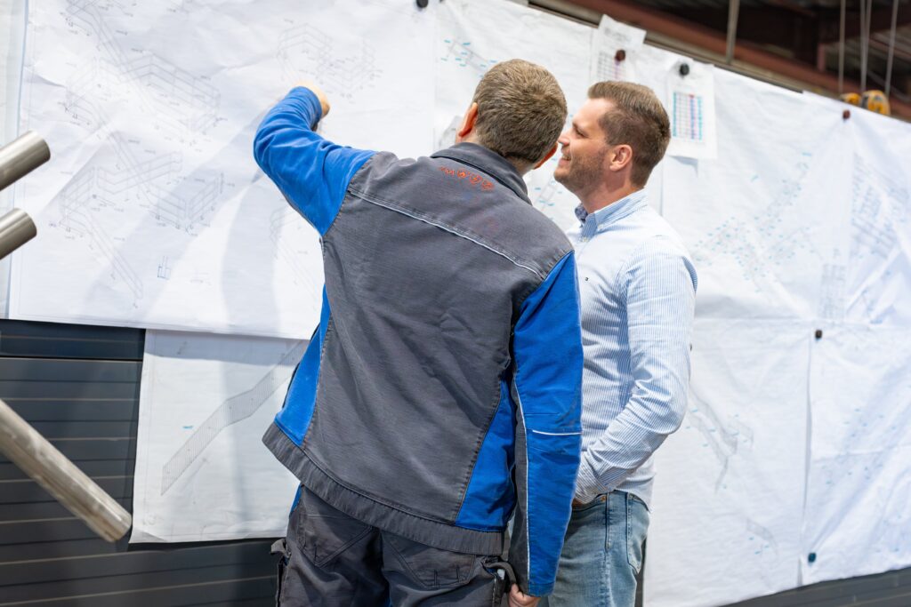 Das Bild zeigt zwei Männer, die intensiv Pläne an einer Wand studieren. Die Wand ist mit großen Papierplänen bedeckt, die technische Zeichnungen und Details zu Konstruktionen zeigen, im Bereich Stahl- oder Metallbau. Der Mann links trägt eine graue Arbeitsjacke mit blauen Ärmeln und zeigt auf einen spezifischen Punkt auf dem Plan, während er dem anderen Mann etwas erklärt. Der Mann rechts trägt ein hellblau gestreiftes Hemd und hört aufmerksam zu. Er scheint in die Erläuterungen vertieft zu sein und blickt konzentriert auf den Plan. Die Szene vermittelt eine professionelle und kollaborative Atmosphäre und zeigt eine typische Arbeitssituation im industriellen Umfeld, in der Pläne und Details gemeinsam besprochen werden, um Präzision und Qualität sicherzustellen.