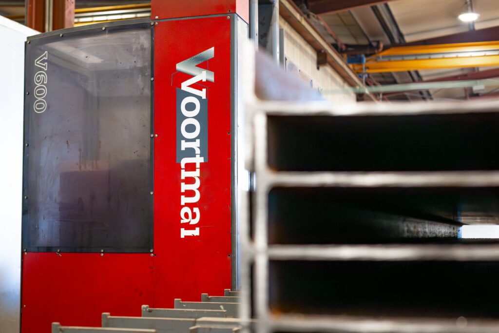 Das Bild zeigt eine rote Industriemaschine der Marke Voortman, Modellnummer V600, die in einer Produktionshalle steht. Die Maschine ist groß und robust und wird für die Bearbeitung von Metallteilen verwendet, was auf die industrielle Ausrichtung des Unternehmens hindeutet. Der Name "Voortman" ist in vertikaler Ausrichtung gut sichtbar auf der Maschine angebracht. Im Vordergrund befindet sich ein unscharfes Metallprofil, das andeutet, dass die Maschine in einem Bereich steht, in dem Stahl- oder Metallträger verarbeitet werden. Die Werkshalle im Hintergrund ist teilweise sichtbar, mit Stahlträgern und einer hohen Decke, die auf eine große Produktionsumgebung schließen lässt. Die Maschine und das Umfeld vermitteln ein Bild von Präzision und industrieller Leistungsfähigkeit und verdeutlichen die Spezialisierung auf Stahlbau und Metallverarbeitung.