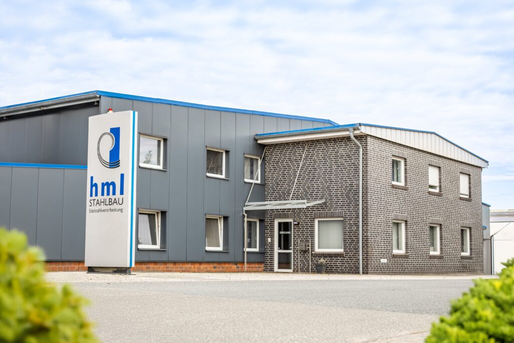 Das Bild zeigt das Firmengebäude der Metallbau HML GmbH in der Siemensstraße 1, 49846 Hoogstede. Das Gebäude besteht aus einer Kombination aus modernen und traditionellen Baumaterialien. Die linke Seite des Gebäudes hat eine graue Metallfassade mit einem blauen Akzent am Dachrand, was auf die industrielle Ausrichtung der Firma hindeutet. Rechts daneben befindet sich ein Teil des Gebäudes mit einer Verkleidung aus dunklen Klinkersteinen, was dem Bau eine zeitlose und solide Anmutung verleiht. Vor dem Gebäude steht ein großes Schild mit dem Firmenlogo und dem Schriftzug "hml STAHLBAU – Edelstahlverarbeitung", das auf die Spezialisierung des Unternehmens hinweist. Der Eingang des Gebäudes ist mit einem kleinen Vordach überdacht und beschildert. Die Umgebung ist ordentlich und gepflegt, und das Bild vermittelt ein professionelles und einladendes Erscheinungsbild des Unternehmensstandorts.