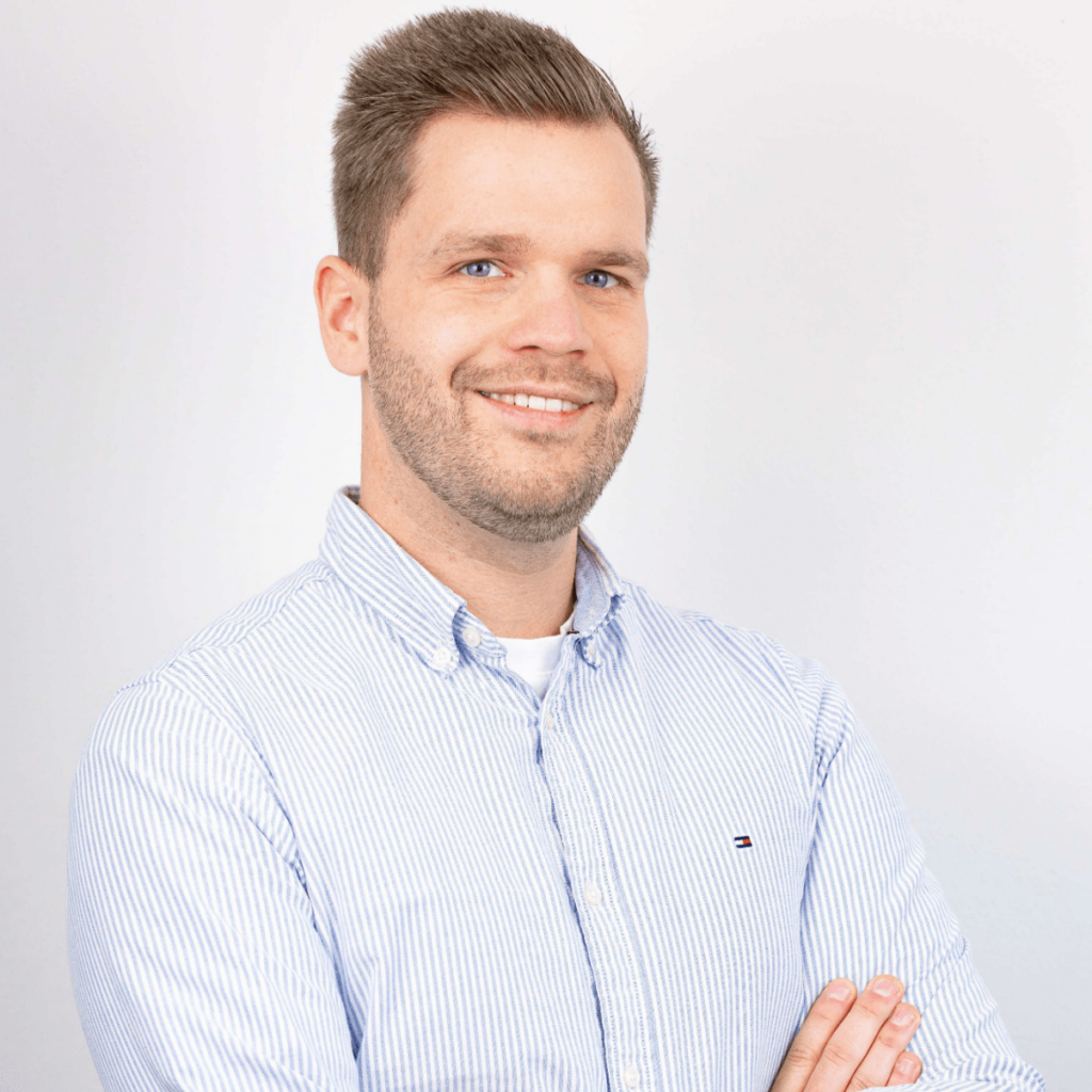 Chris Vahrenhorst: Porträtfoto eines jungen Mannes mit kurzen, braunen Haaren und einem blauen gestreiften Hemd. Er lächelt freundlich und hat ebenfalls die Arme verschränkt. Seine Rolle ist die Projektleitung.