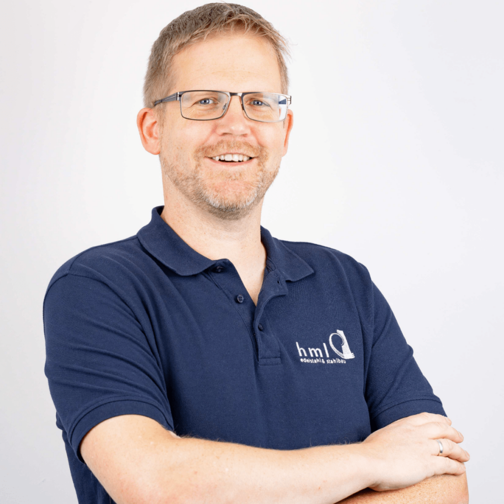 Jörg Westerhoff: Porträtfoto eines Mannes mittleren Alters mit kurzem, blondem Haar und Brille. Er trägt ein dunkelblaues Polohemd mit Firmenlogo und lächelt freundlich in die Kamera, während seine Arme vor der Brust verschränkt sind. Seine Rolle ist Technische Systemplanung.