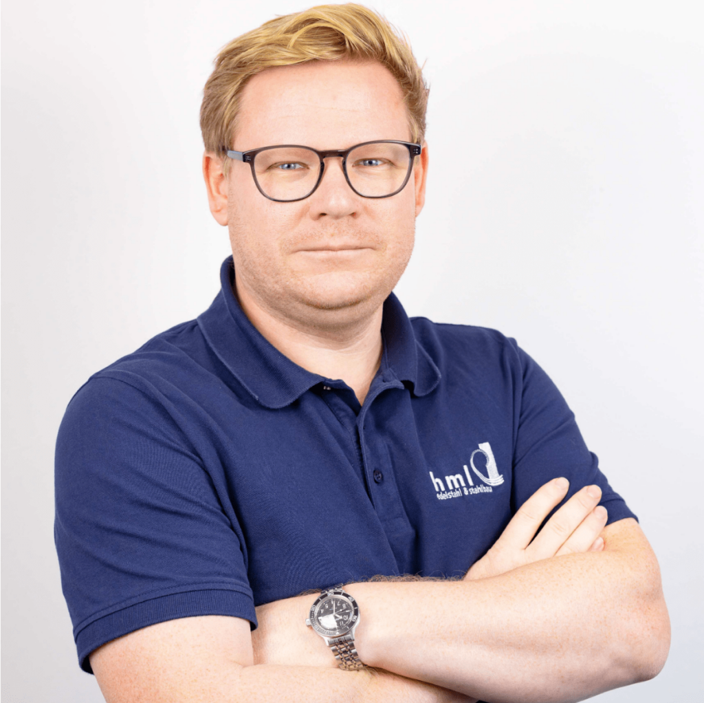 Arthur Kempf: Porträtfoto eines Mannes mittleren Alters mit blondem Haar und Brille, der ein dunkelblaues Polohemd mit Firmenlogo trägt. Seine Arme sind vor der Brust verschränkt, und er wirkt konzentriert. Seine Rolle ist Schweißfachingenieur und verantwortlich für die Kalkulation im Hallenbau.